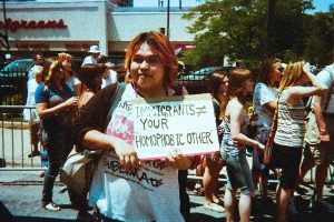 プライドパレードで手書きのプラカードを持つマサキ。プラカードには "IMMIGRANTS ≠ YOUR HOMOPHOBIC OTHER" とある。