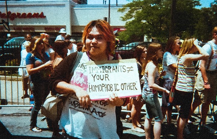 プライドパレードで手書きのプラカードを持つマサキ。プラカードには "IMMIGRANTS ≠ YOUR HOMOPHOBIC OTHER" とある。